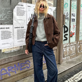 Person wearing a brown jacket and blue jeans standing in front of a wall with posters and graffiti.